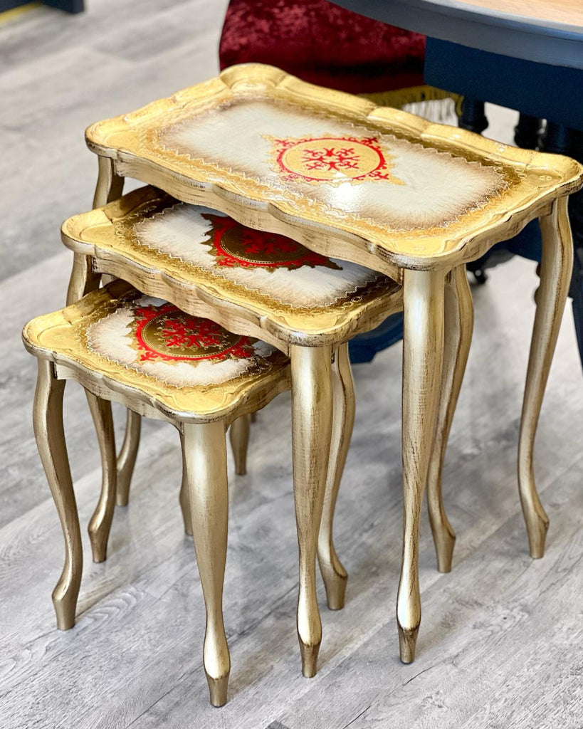 A set of three gold-colored, maroon and white designed nesting tables made of wood, with ornate legs and detailing.
